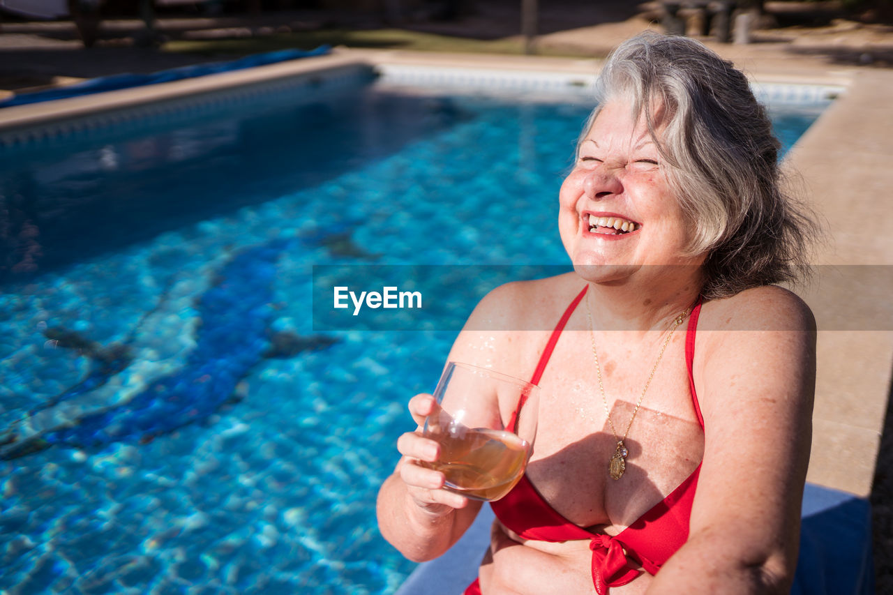 High angle of delighted senior female tourist in bikini laughing brightly while chilling on poolside with drink