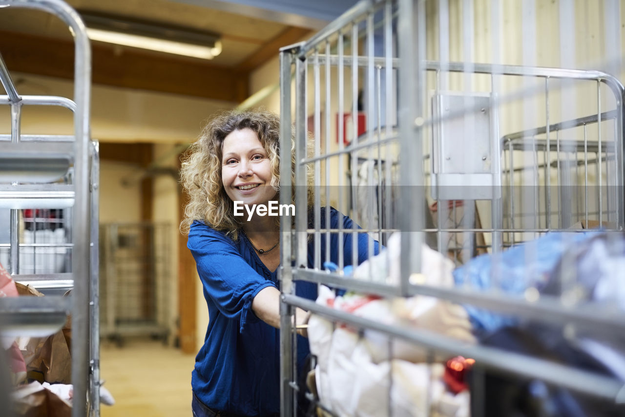 Smiling mature female volunteer pushing cart while looking away at warehouse