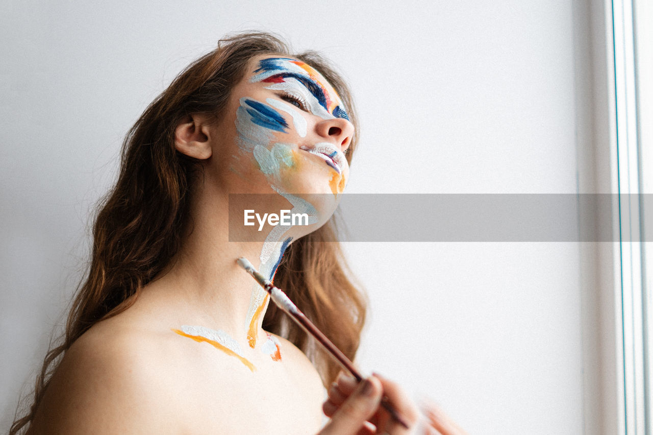 Close-up of young woman applying face paint against wall