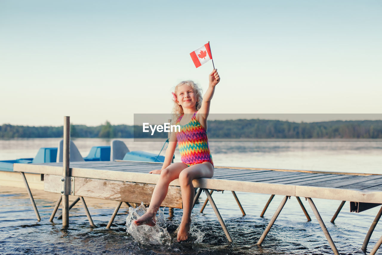 Girl sitting on dock pier by lake and waving canadian flag. canada day holiday on july outdoor.