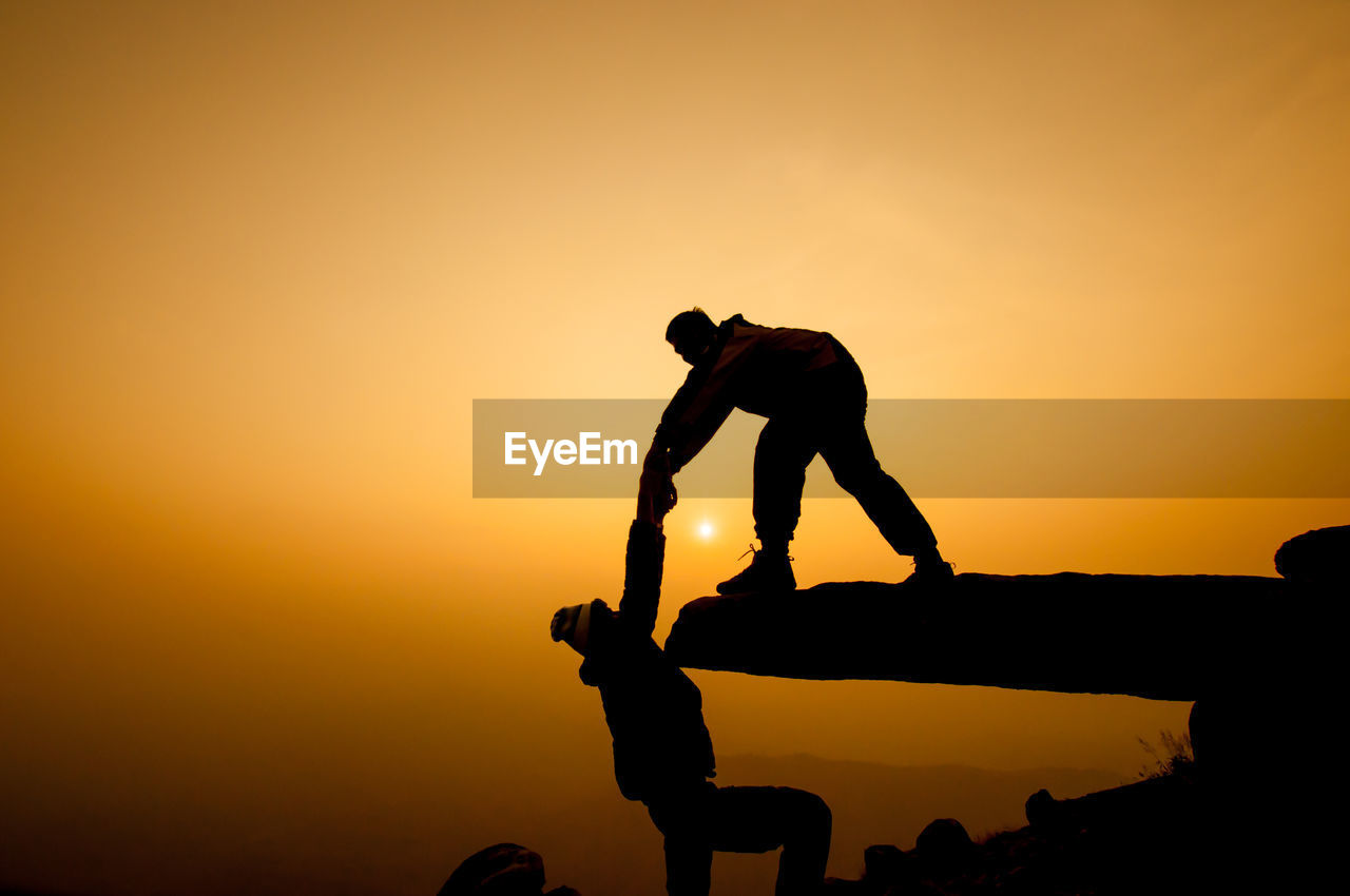 Silhouette man assisting friend climbing cliff against orange sky during sunset