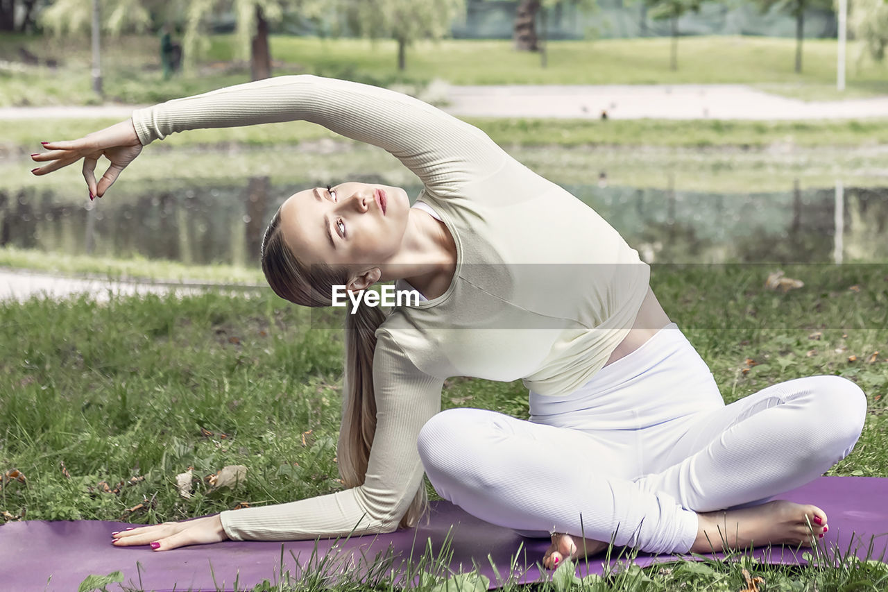 A young woman with long blonde hair does yoga in the summer in nature by the pond in the park.