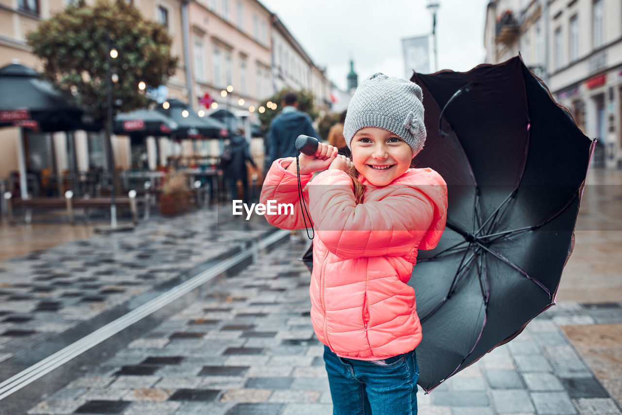 Little smiling happy girl holding big umbrella walking in a downtown on rainy gloomy autumn day