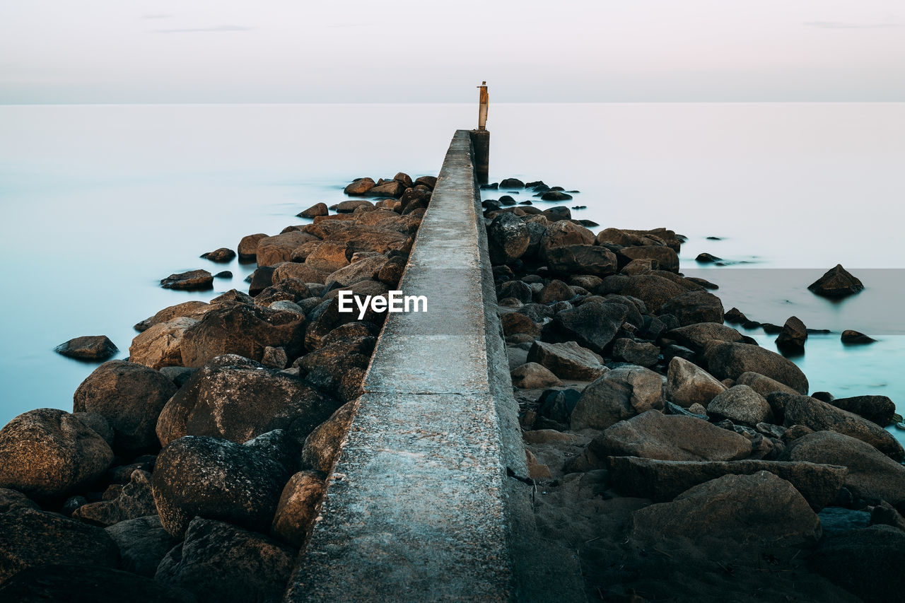 Rocks on beach against sky. pier stretching out in a calm sea. long exposure 