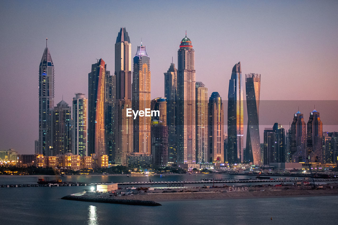 Illuminated modern buildings by bay against sky