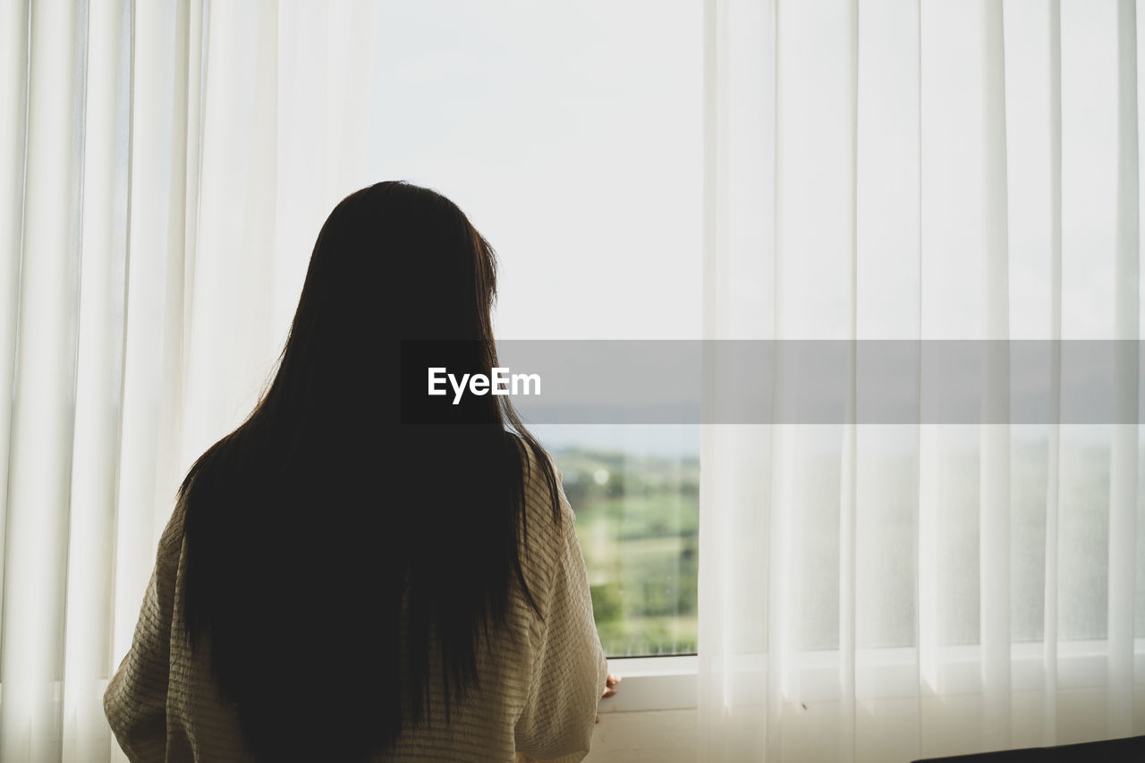 one person, window, indoors, adult, women, long hair, white, black, hairstyle, rear view, curtain, day, young adult, waist up, sadness, lifestyles, loneliness, contemplation, looking, looking through window, person, dress, depression - sadness, window covering, standing, window treatment, copy space, home interior, emotion, sitting, interior design, casual clothing, solitude, sunlight, nature