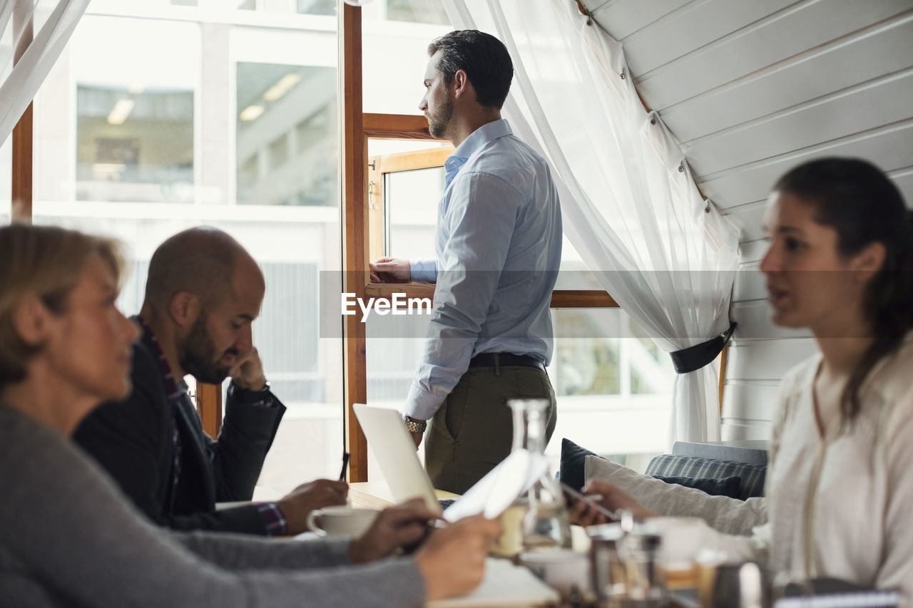 Businessman looking through office window while colleagues discussing in foreground