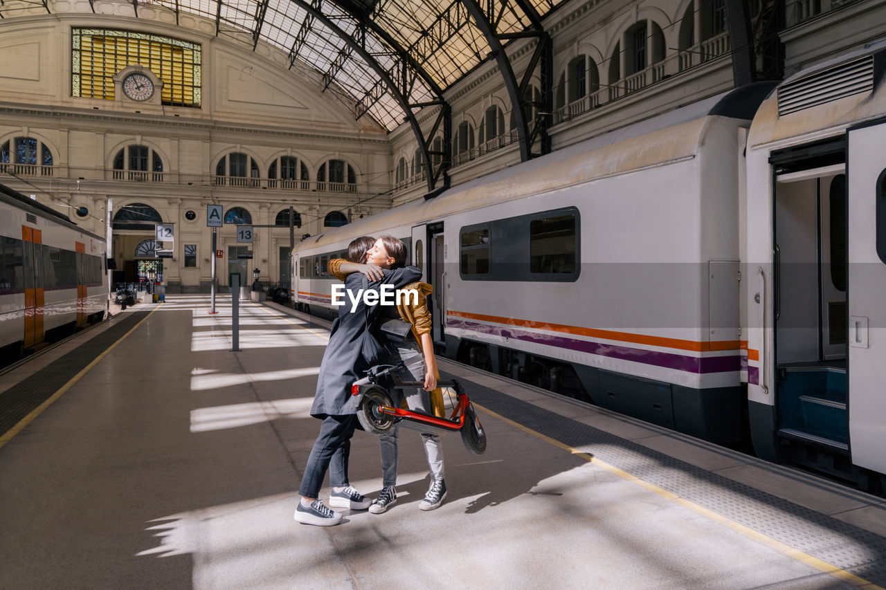 Young woman holding push scooter embracing friend at railroad station