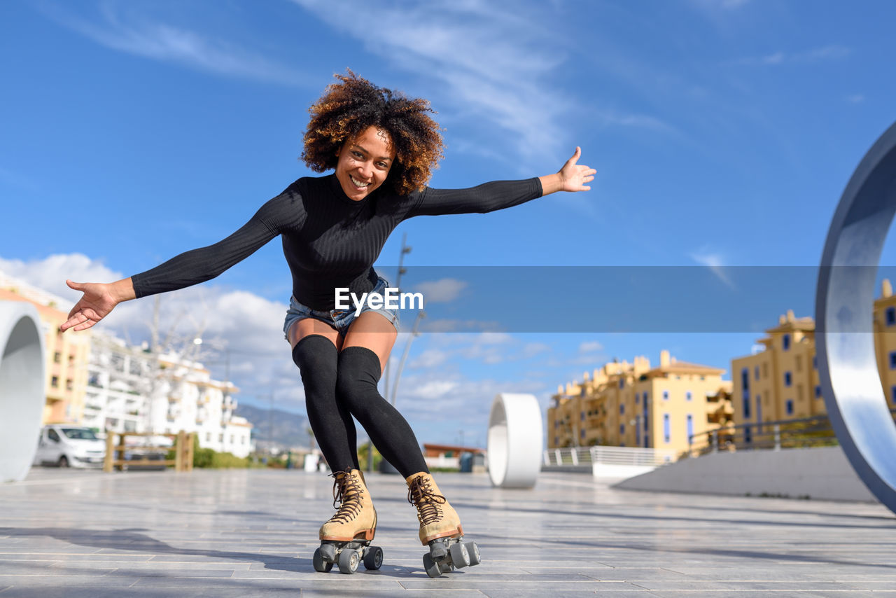 Portrait of mid adult woman with arms outstretched roller skating against blue sky in city