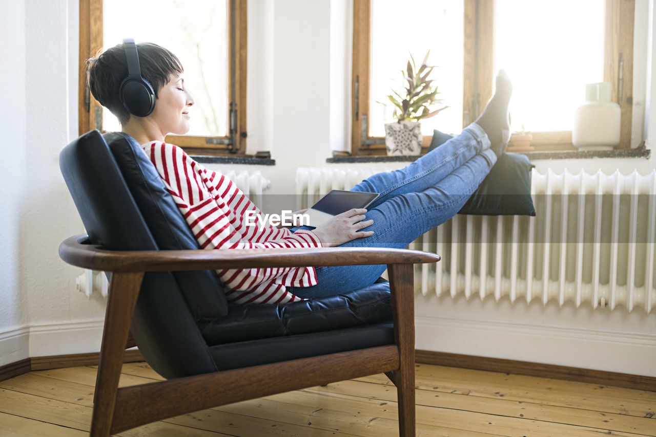 Short-haired woman with headphones relaxing in lounge chair in stylish apartment