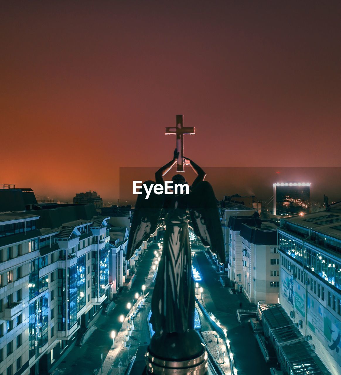 Angel statue with cross against sky in city at night