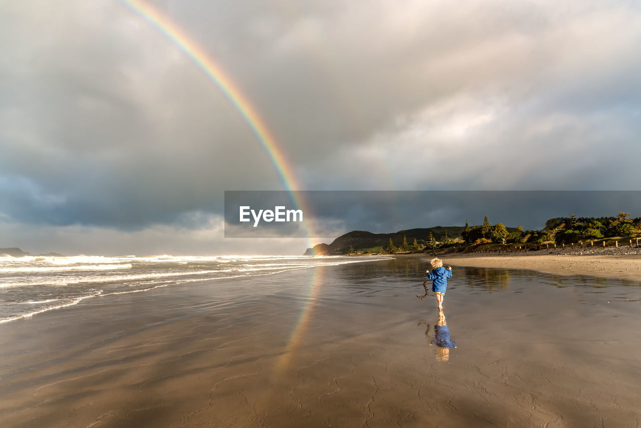 Toddler holding seaweed playing on beach with rainbow and reflection