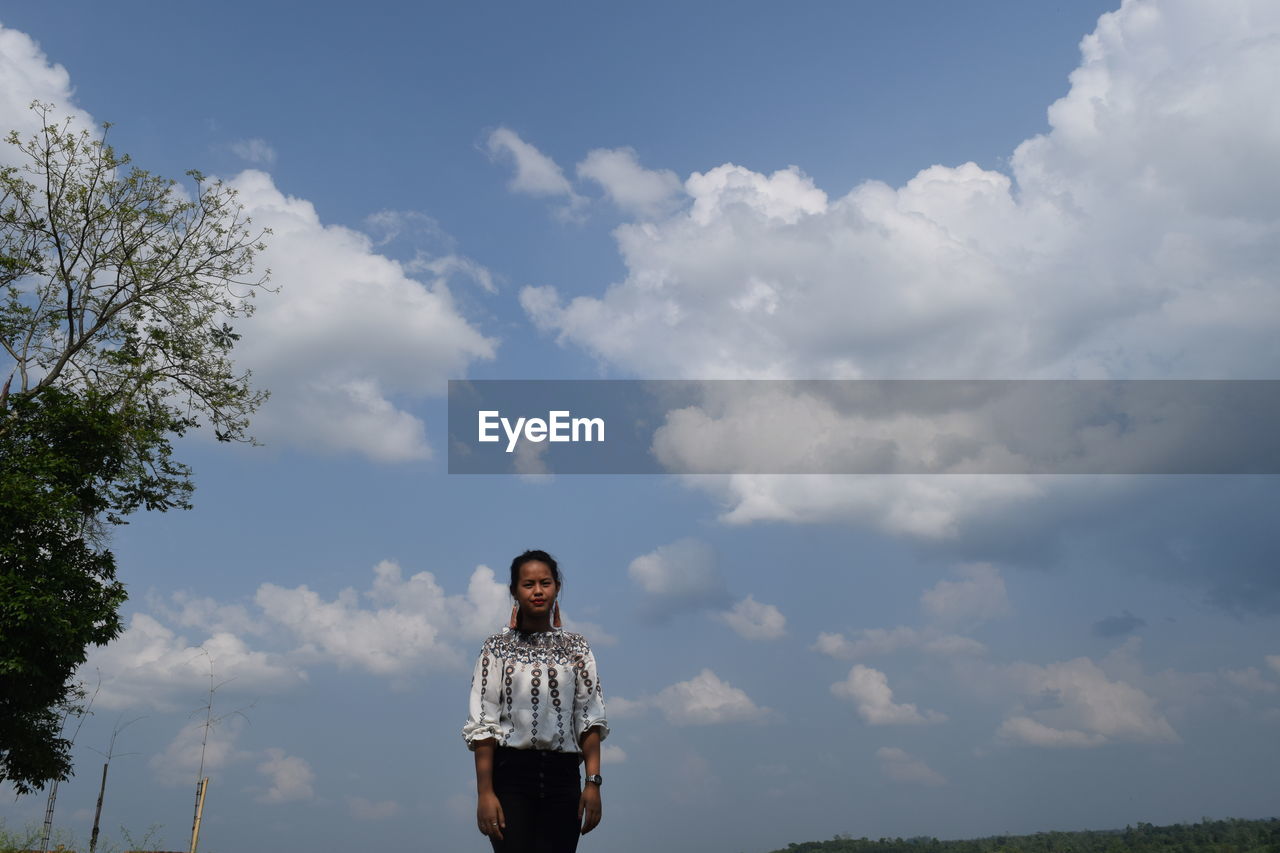 Portrait of young woman standing against cloudy sky