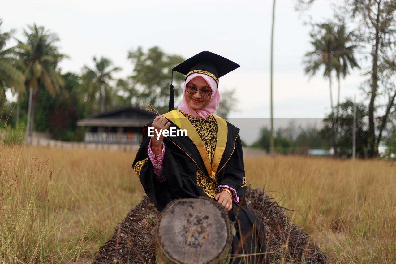 Portrait of woman in graduation gown sitting on log