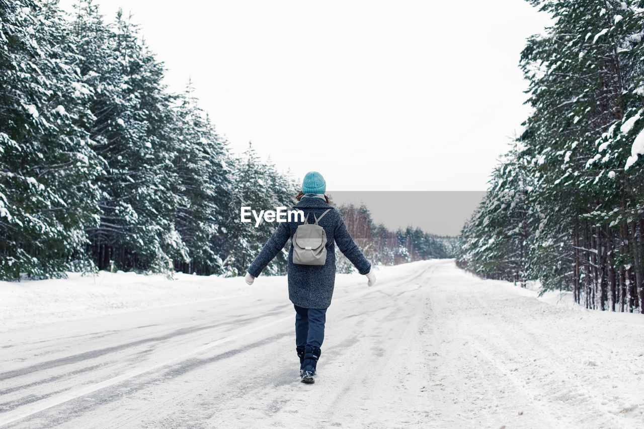 A girl in a coat and a hat, with a  backpack on her back, walks along the side of a winter road