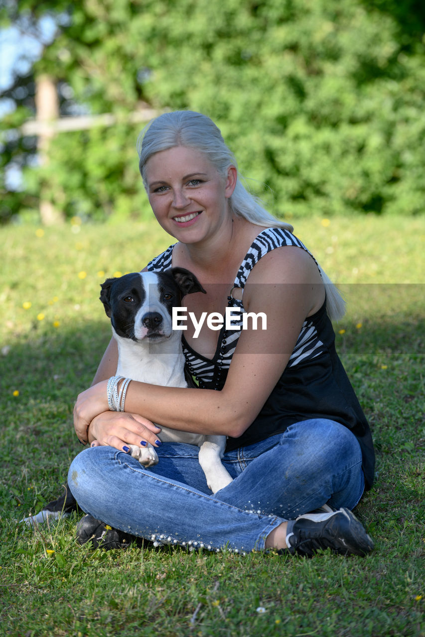 A young blonde woman is sitting on a freshly green lawn and hugging and cuddling her dog.