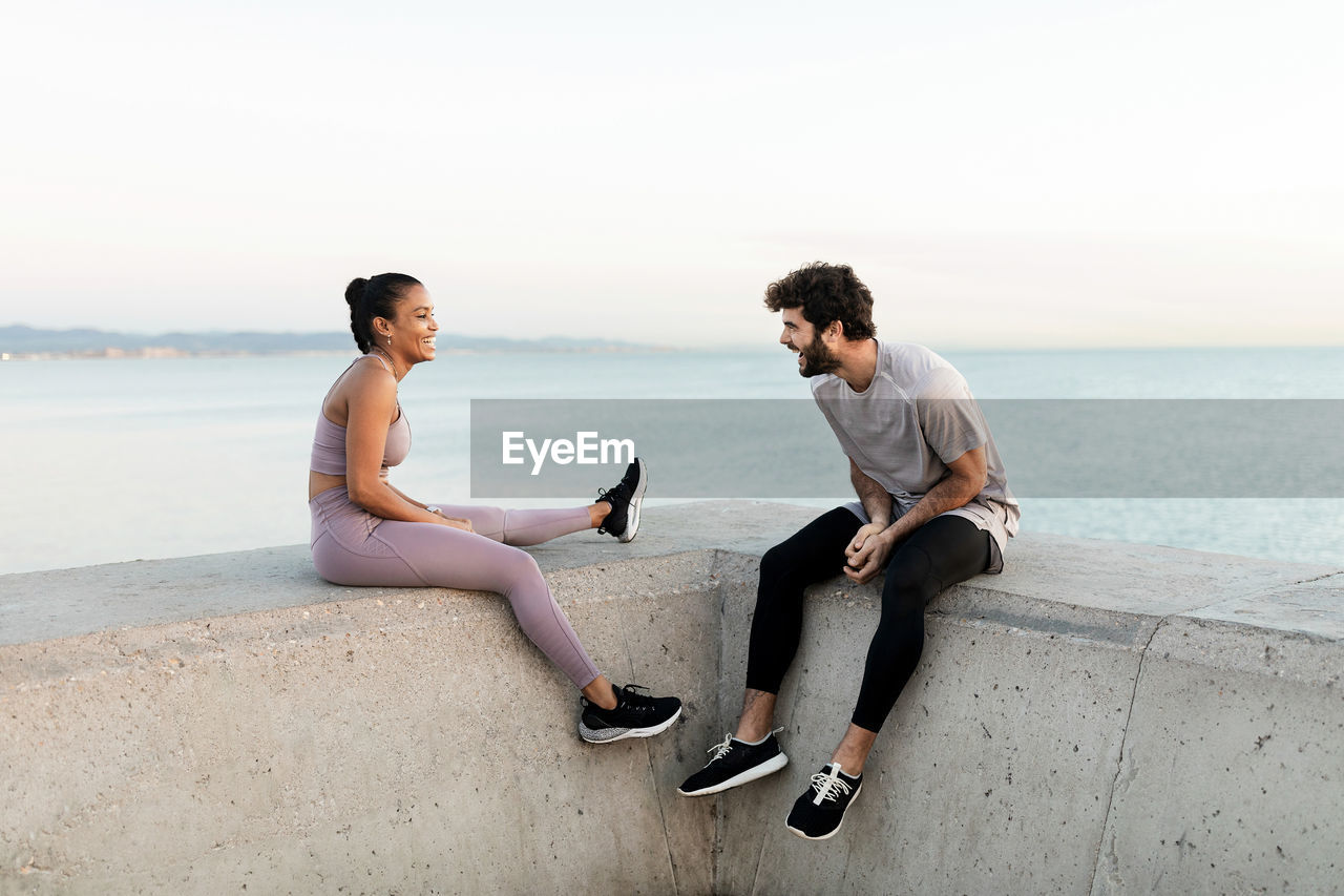 Cheerful young bearded sportsman speaking with content ethnic female athlete while resting against endless sea and looking at each other