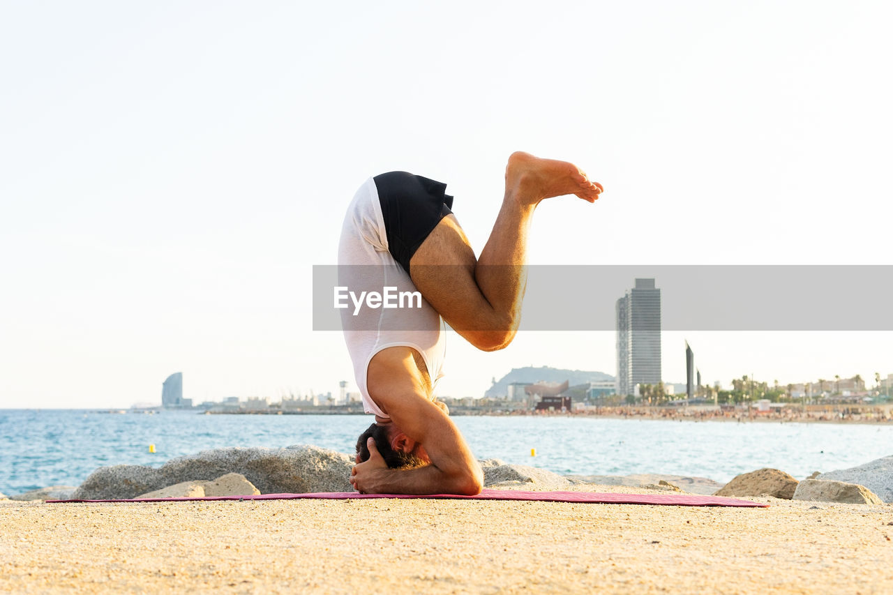 Full body unrecognizable barefoot male doing salamba sirsasana pose on beach near sea during yoga session in morning