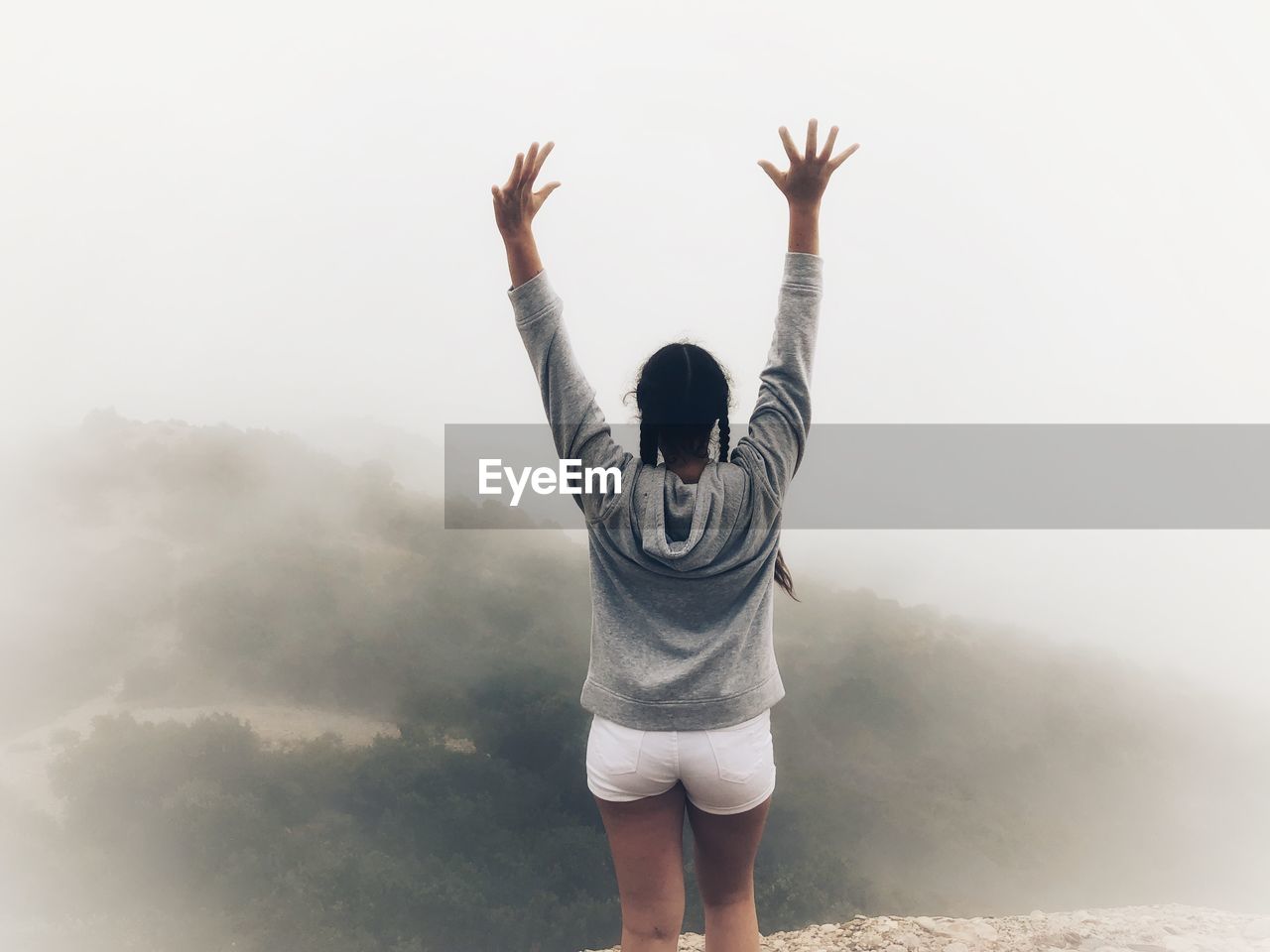 Rear view of teenage girl with arms raised standing on mountain during foggy weather