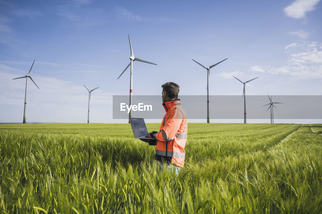 Engineer with laptop on wheat field looking at wind turbines