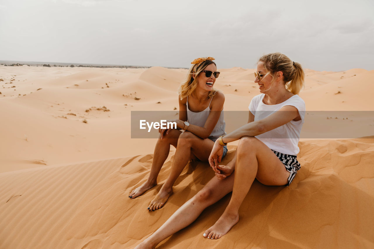 Cheerful friends sitting on sand at desert against sky