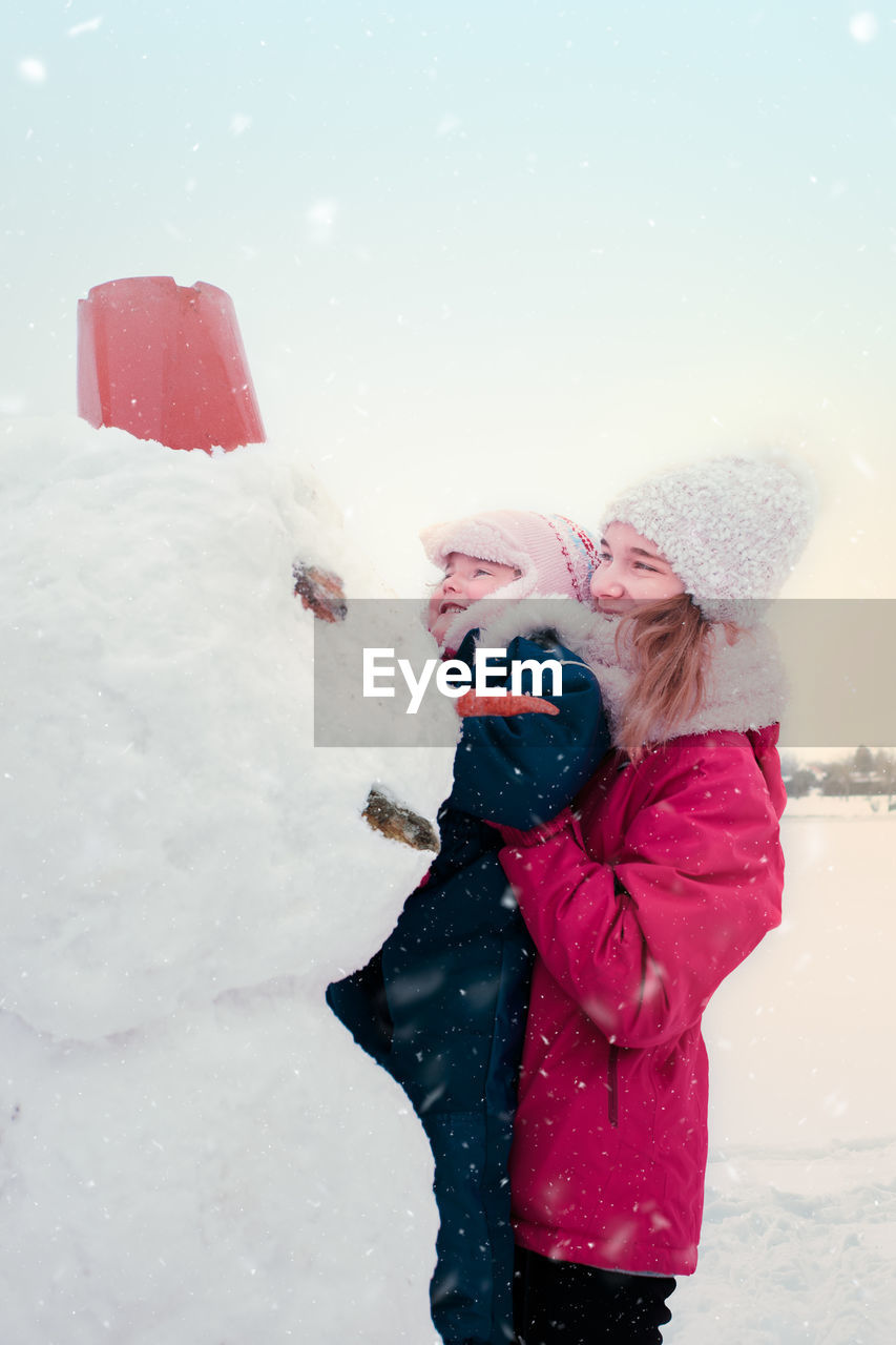 Teenage girl carrying sister by snowman against sky