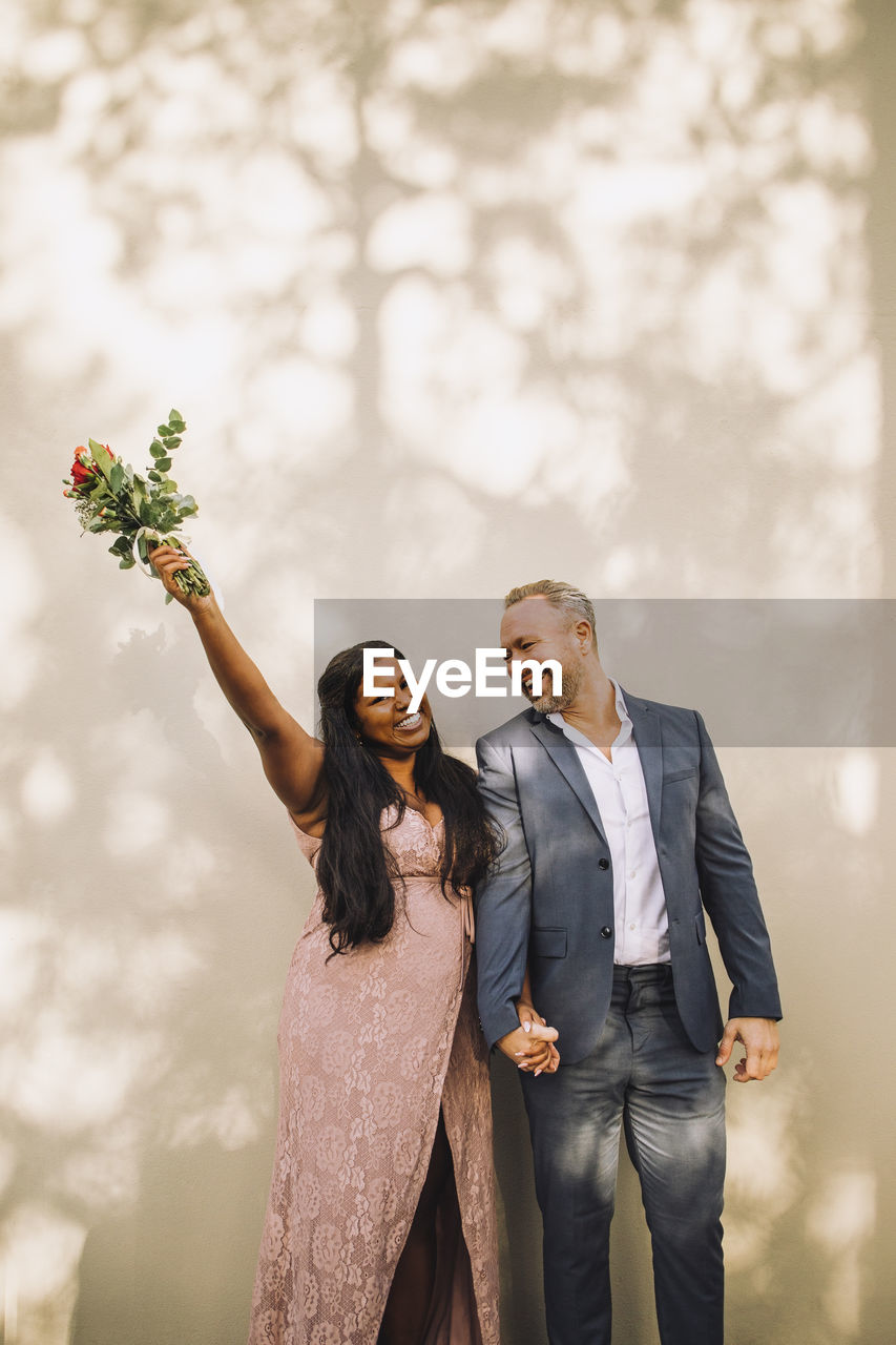 Happy bride holding bouquet with hand raised standing by groom against wall