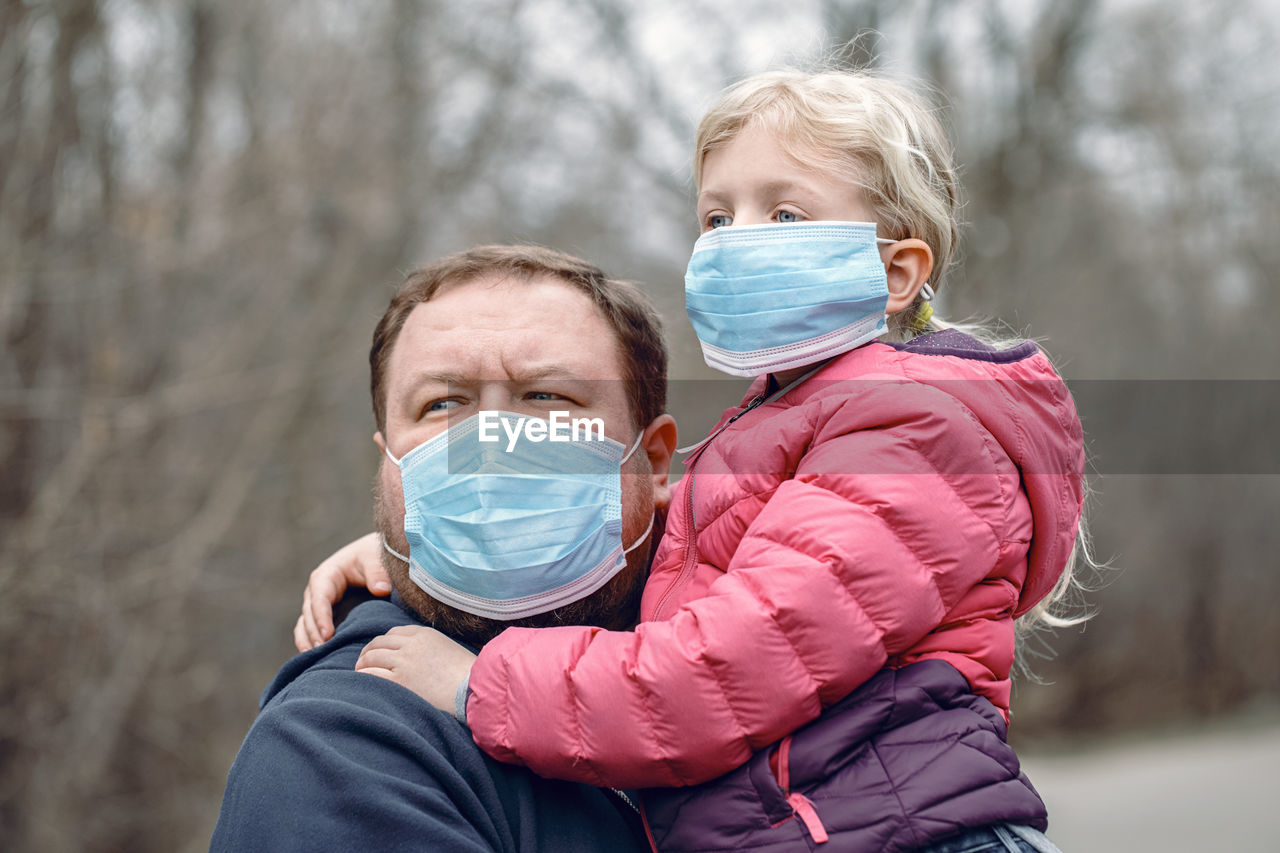 Father and daughter wearing flu mask looking away standing outdoors