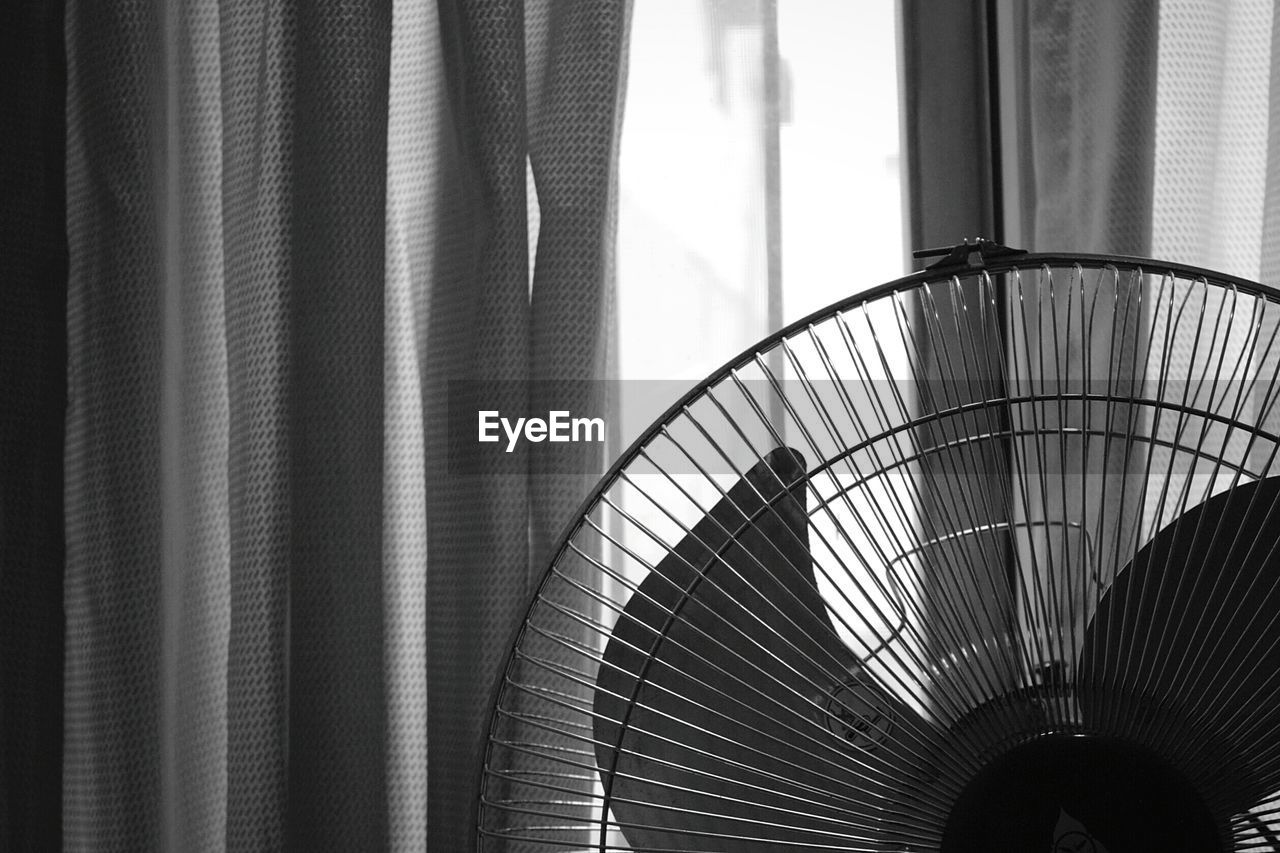 Close-up of cropped electric fan against curtained window