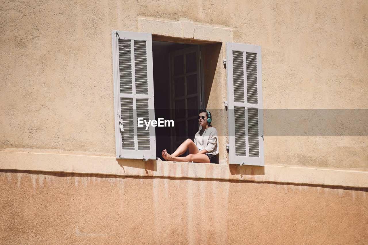 Low angle view of woman hearing music while sitting on window