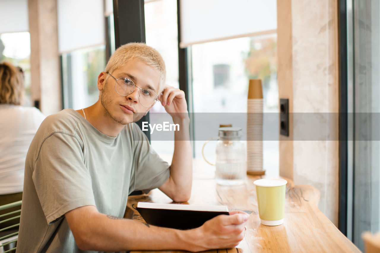 Stylish blond teenage boy 18-19 year old wear glasses reading paper book in cafe with coffee in cup.