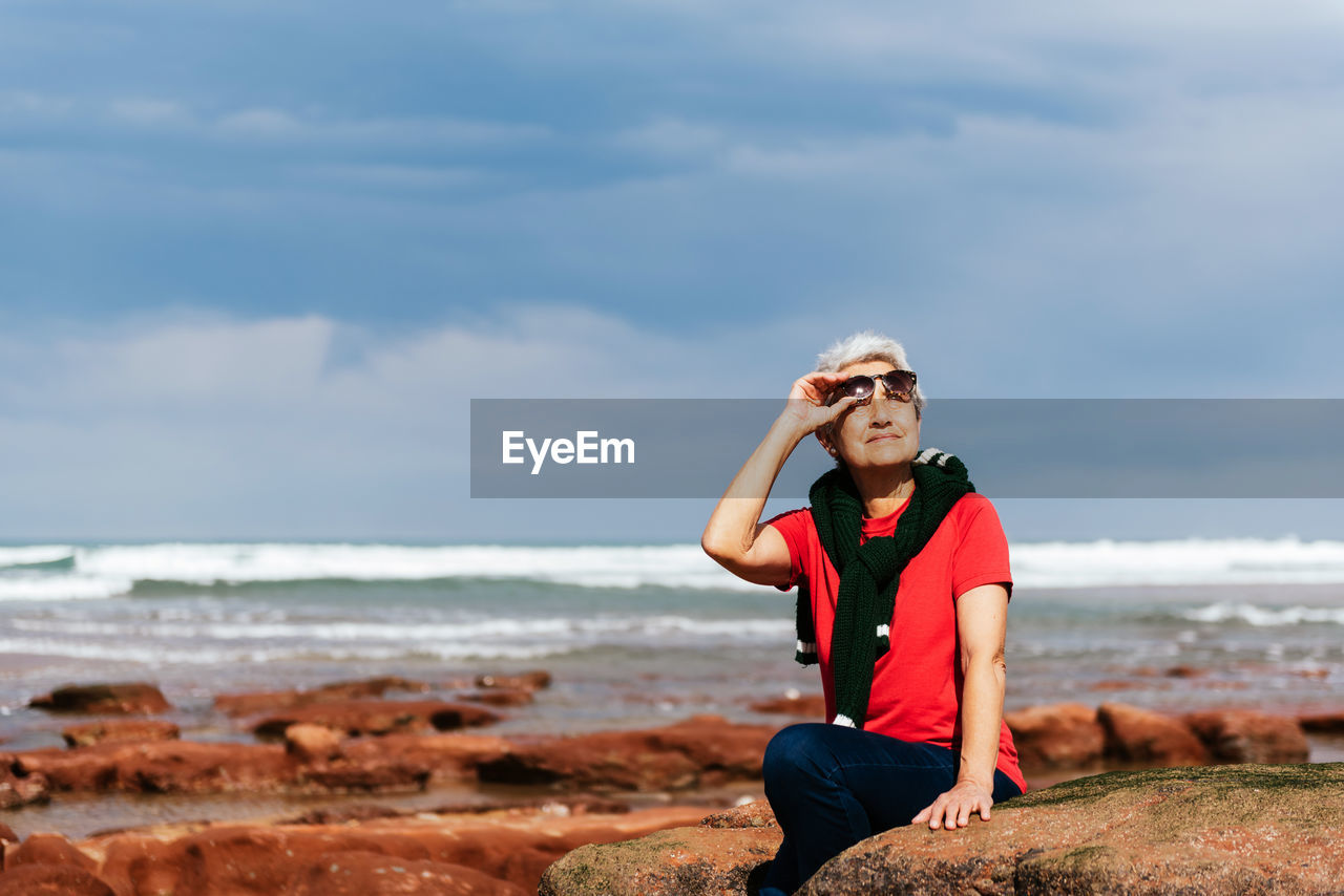 Smiling elderly female tourist in sunglasses sitting on rough boulder while looking up against ocean under cloudy sky