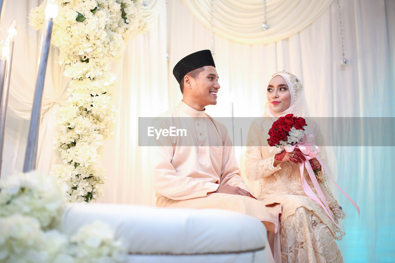 Bride holding flower bouquet by groom sitting at home