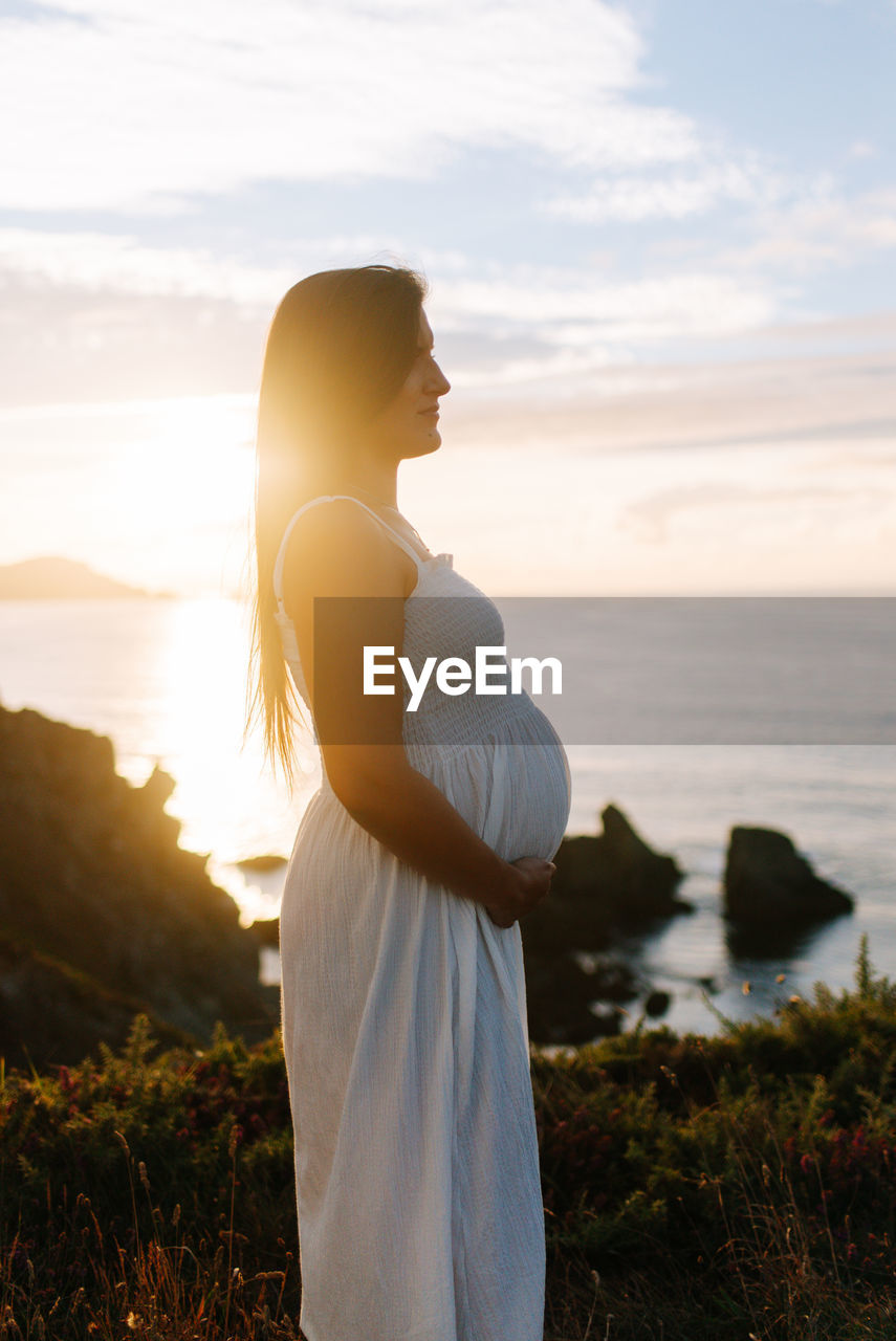 Side view of pregnant woman standing against sky during sunset