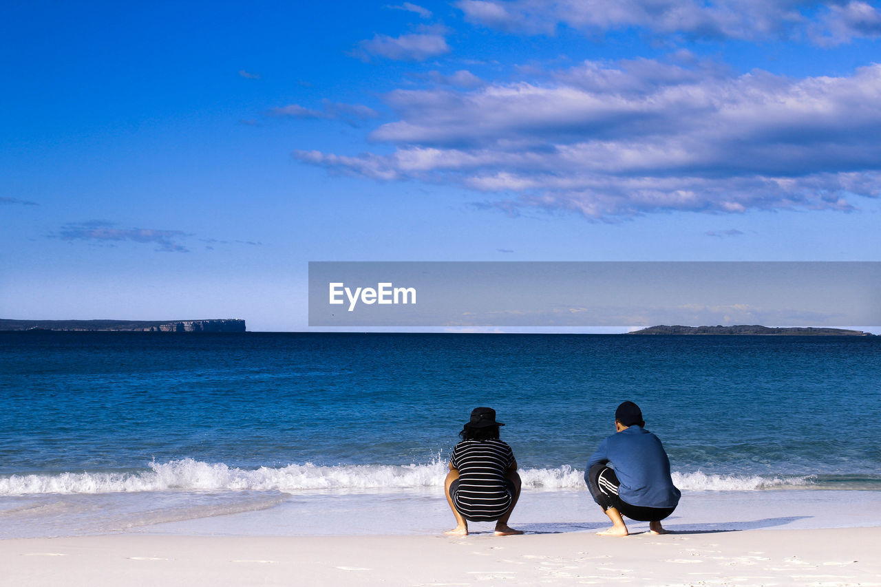 Rear view of men crouching at beach against clear blue sky with some clouds