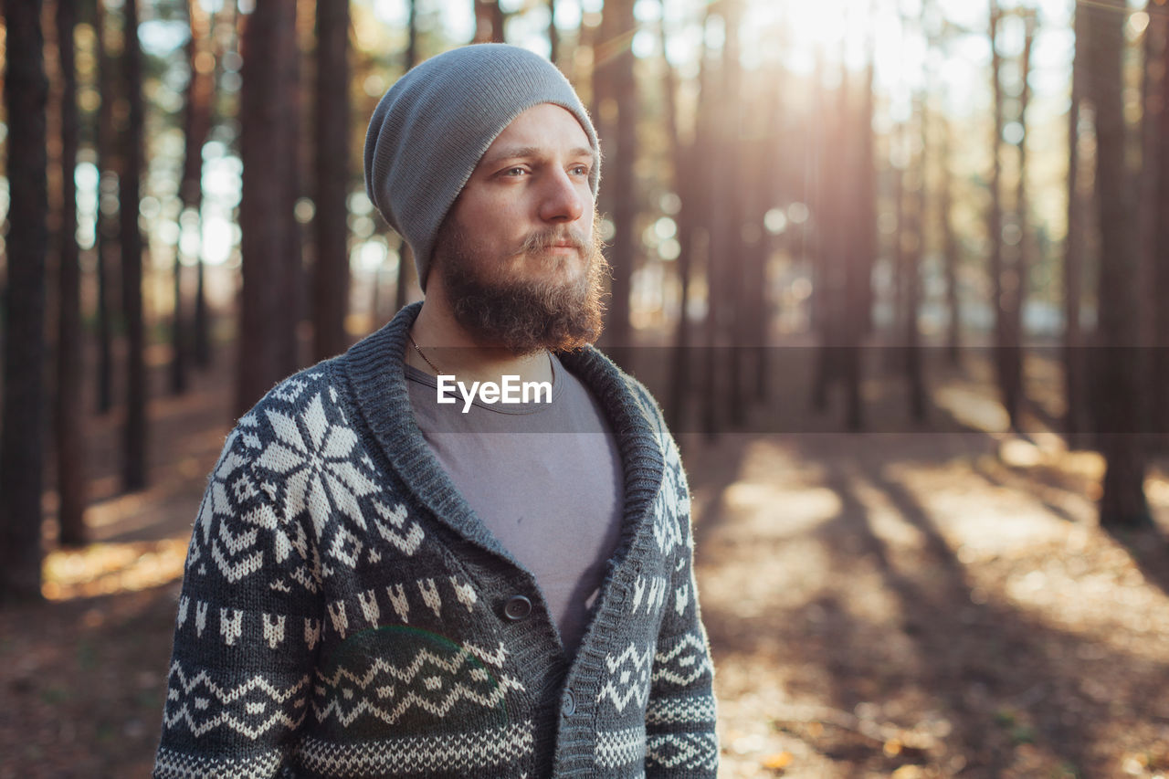 Hipster looking away while standing against trees in forest