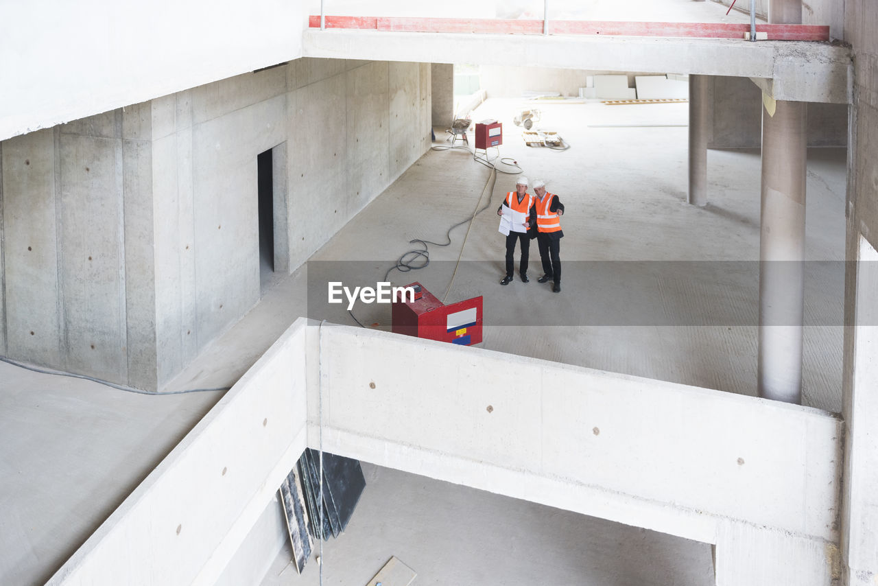 Two men with plan wearing safety vests talking in building under construction