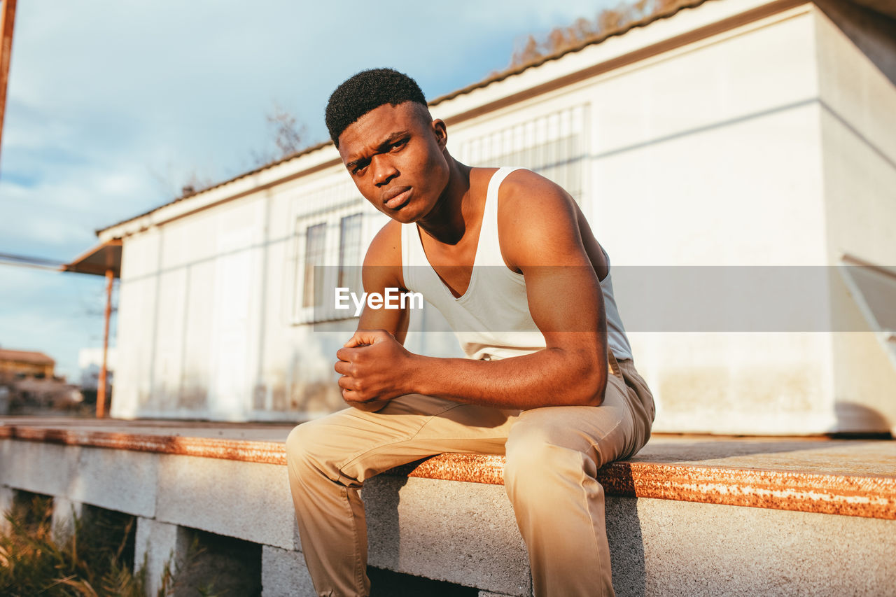 Young masculine african american male in undershirt with clasped hands looking at camera against house