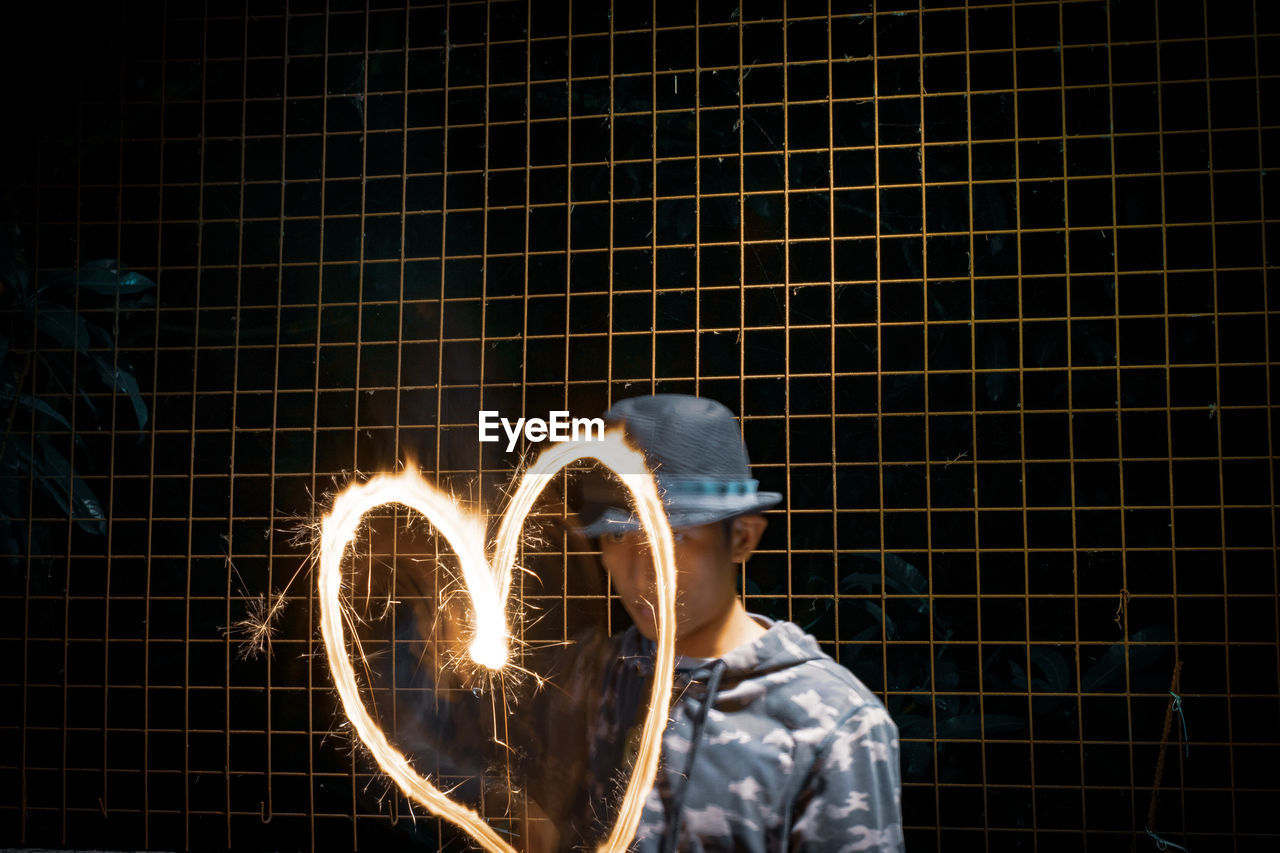 Man making heart shape with sparkler while standing against metal fence