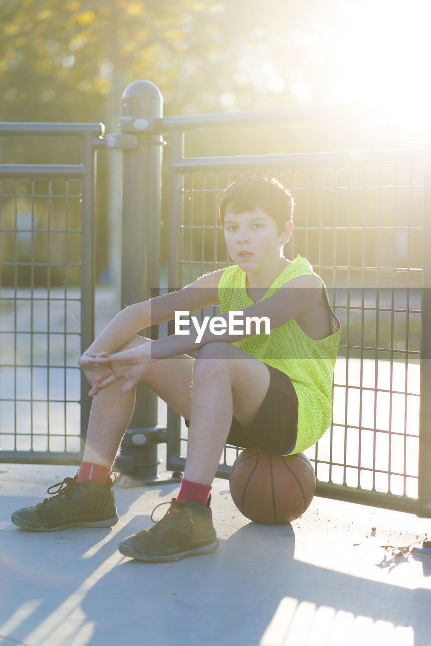 Portrait of boy sitting on basketball against fence
