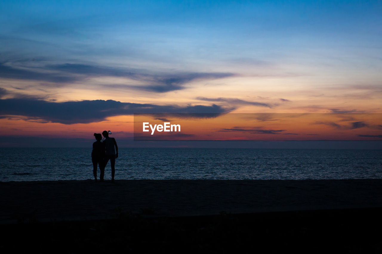 SILHOUETTE COUPLE STANDING ON BEACH AGAINST SKY DURING SUNSET