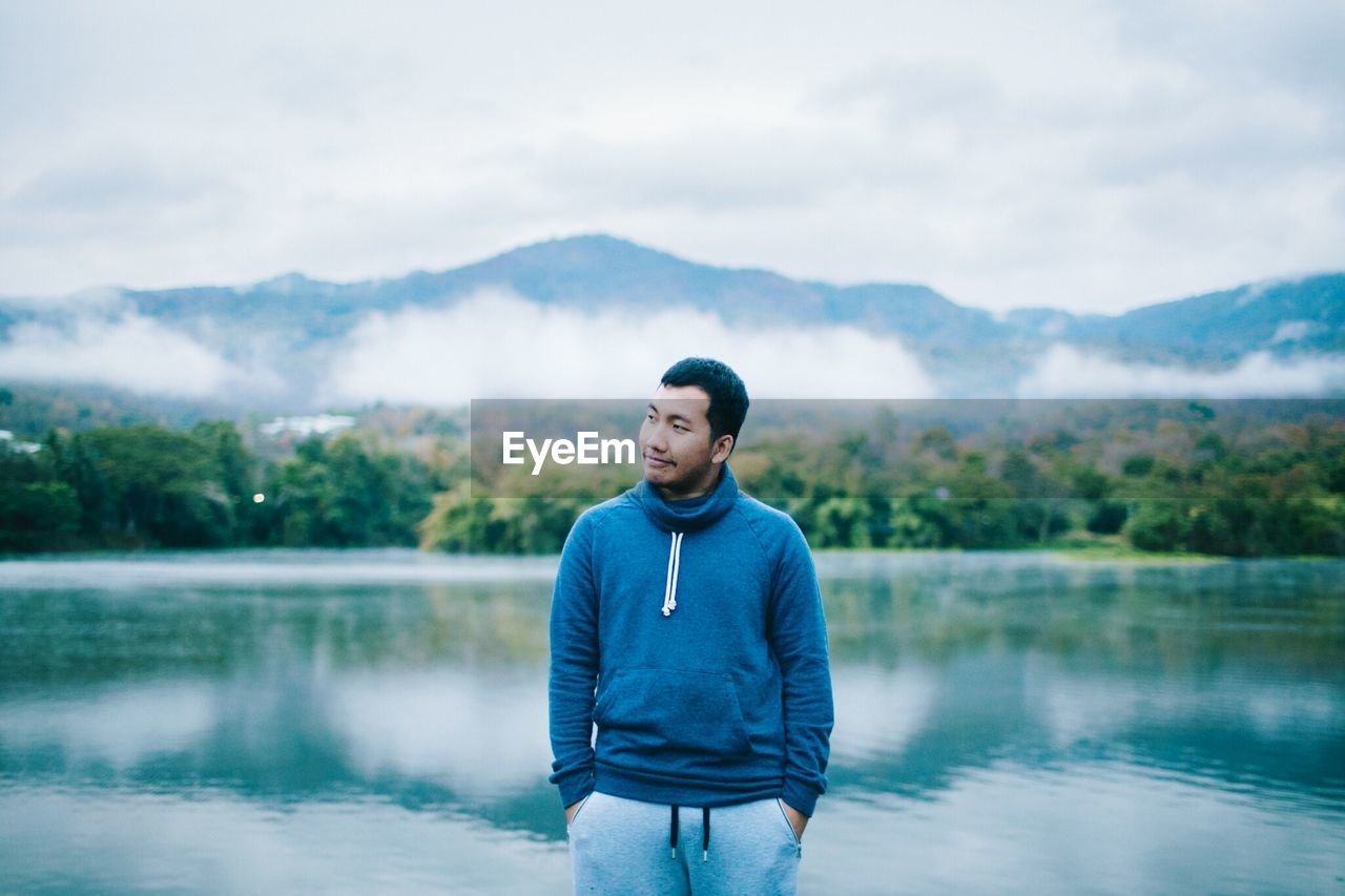 MAN STANDING IN FRONT OF LAKE AGAINST MOUNTAINS