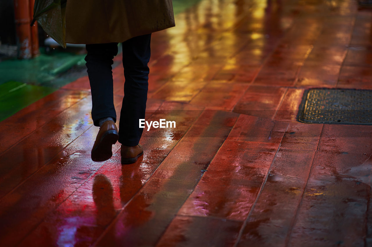 Low section of man walking on wet footpath in city at night