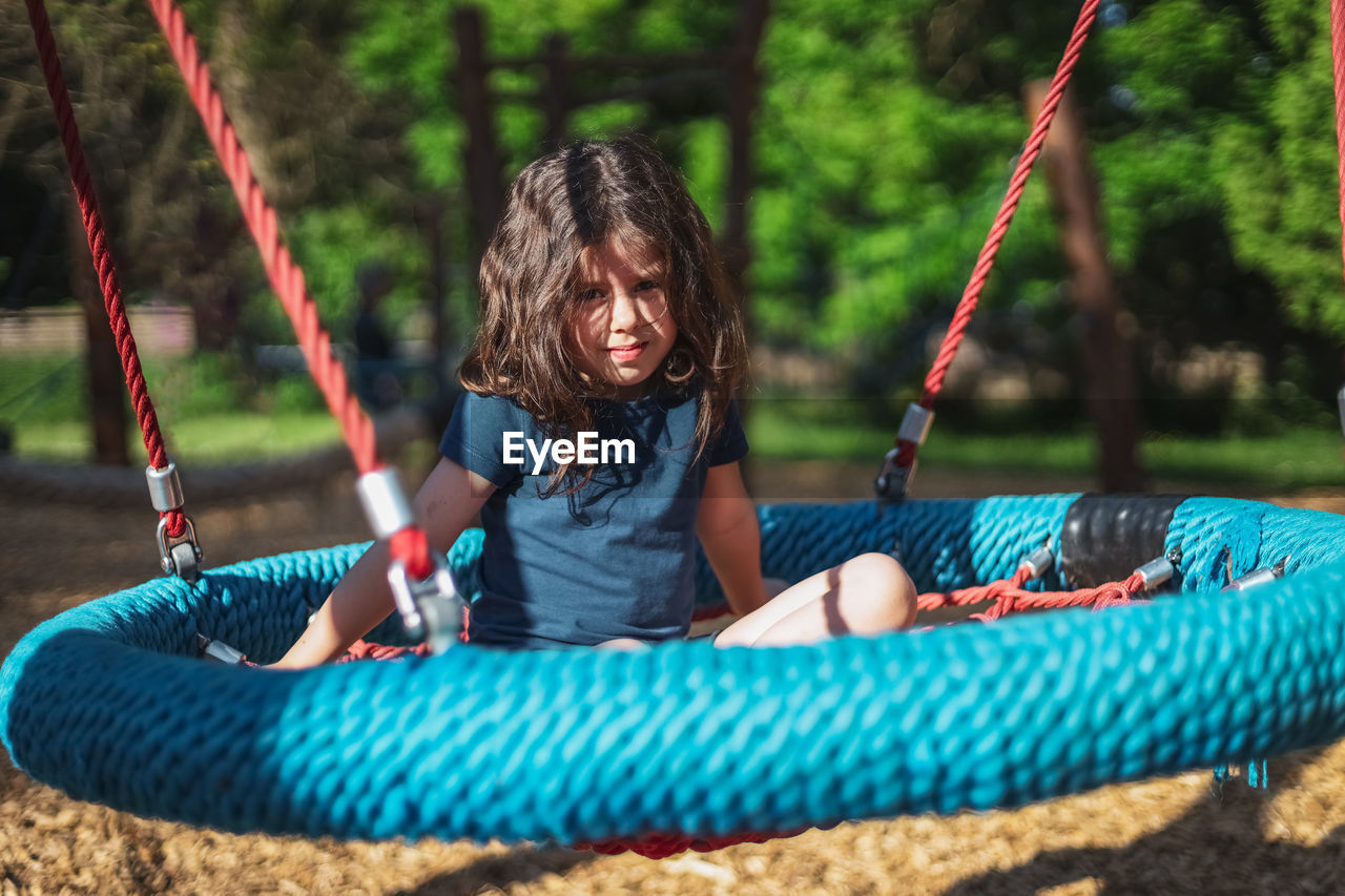 Portrait of a beautiful caucasian girl sits on a round rope swing in the park at the playground