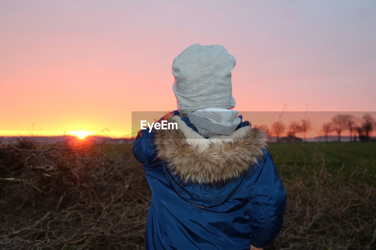 Rear view of kid standing on field against sky during sunset