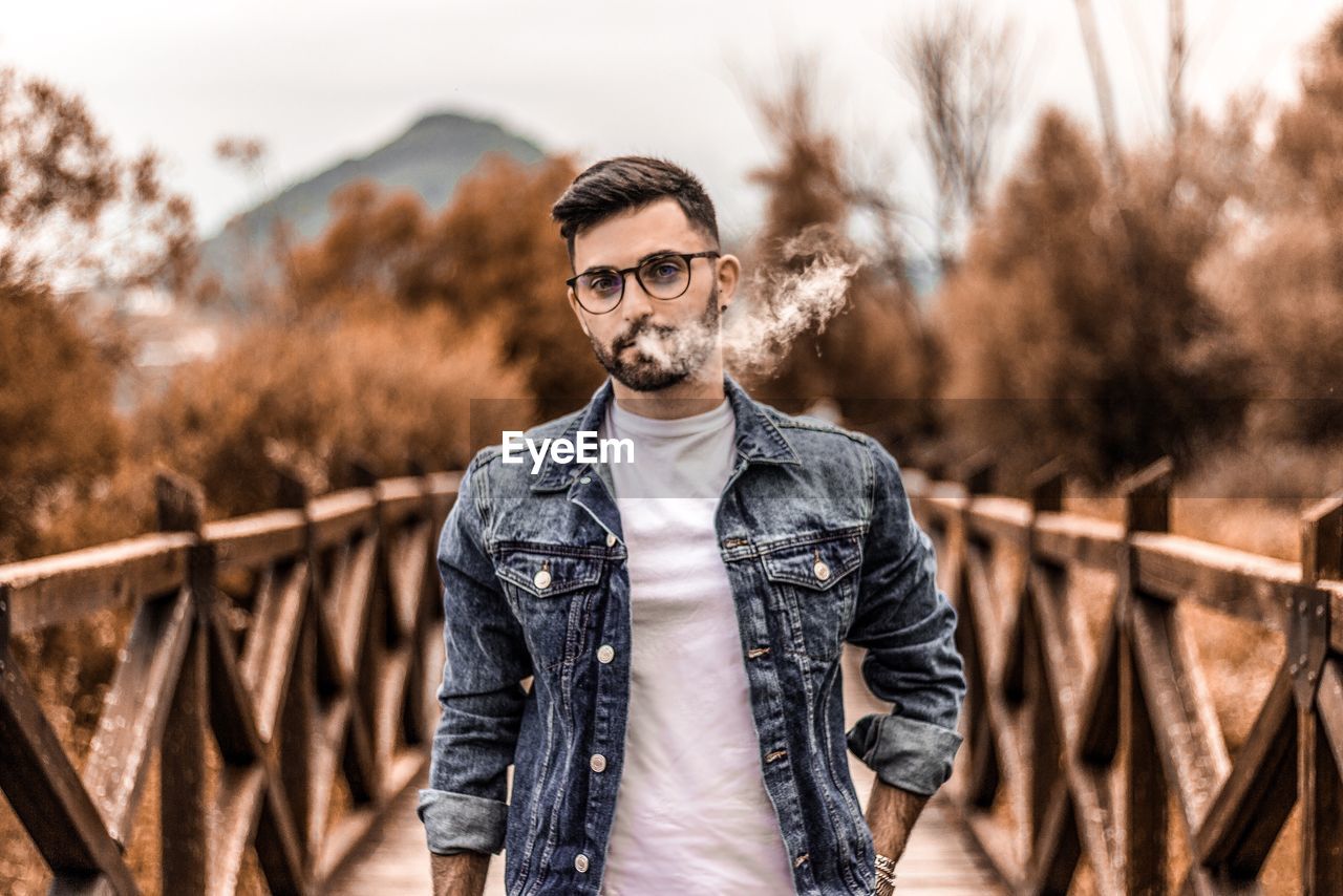 Portrait of young man smoking while standing on footpath against trees