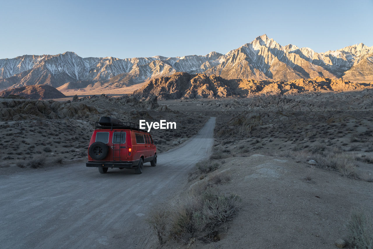 Red camper vehicle driving between fields on long roadway against range of rough rocky cliffs covered with snow on winter day in alabama hills, california, usa