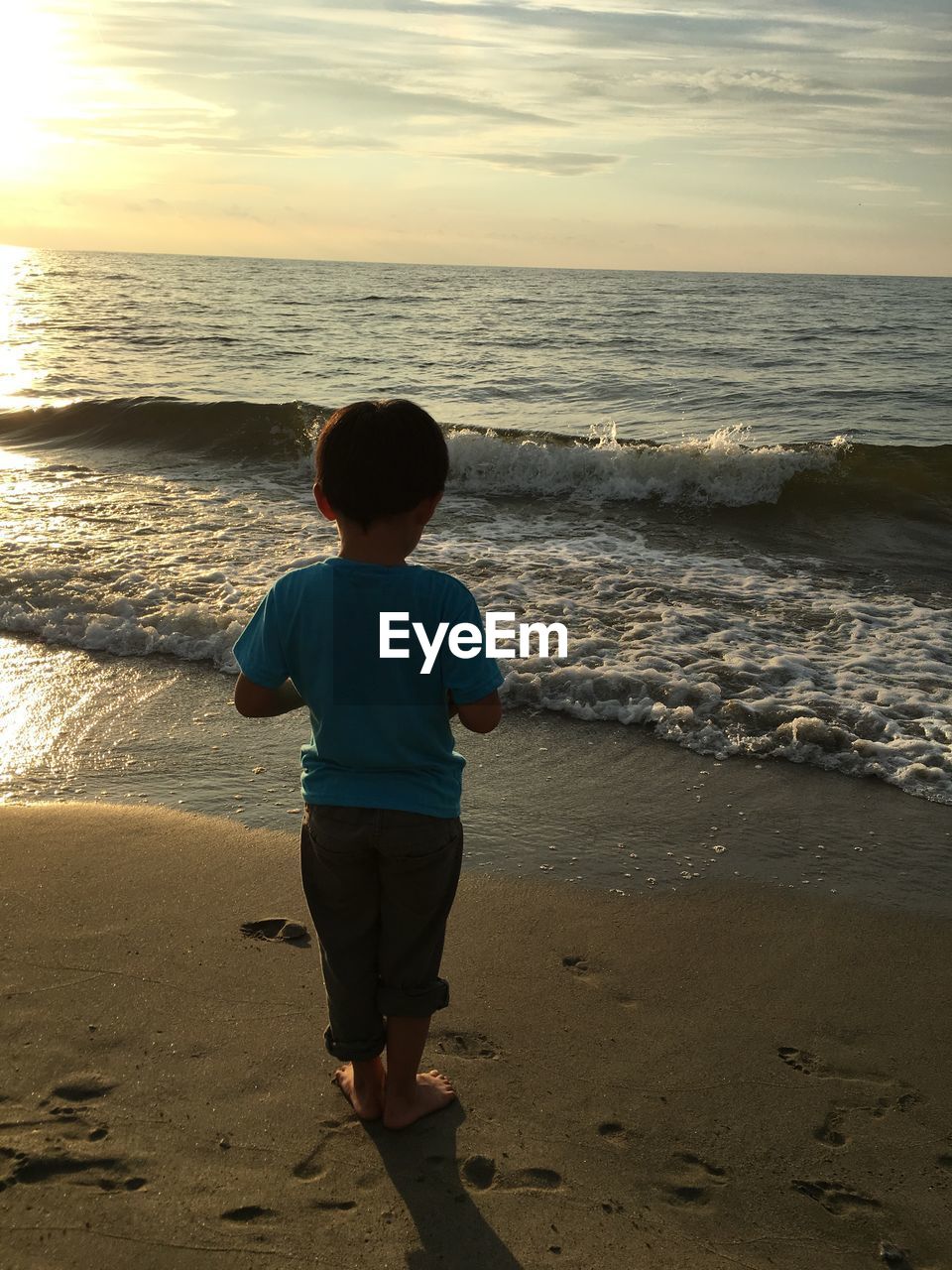 Rear view of boy standing on shore at beach during sunset