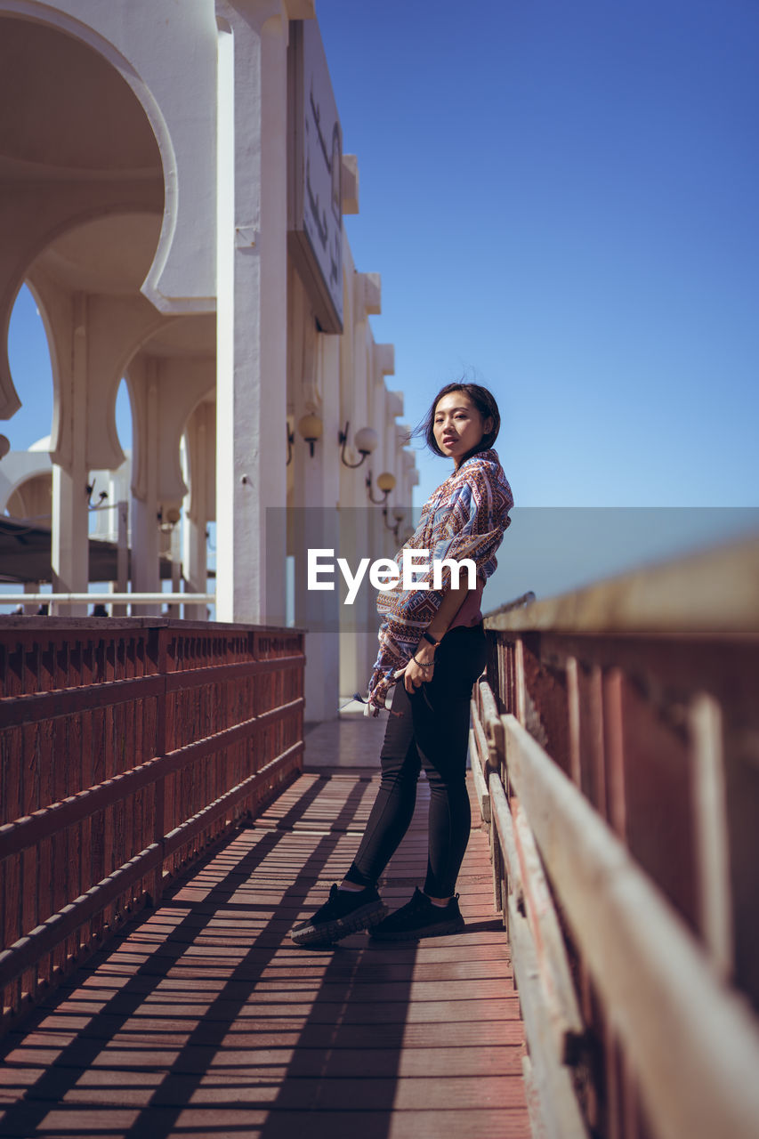 Positive young asian female in colorful traditional headscarf smiling and looking at camera while standing near beautiful white building of al rahma mosque in jeddah in saudi arabia