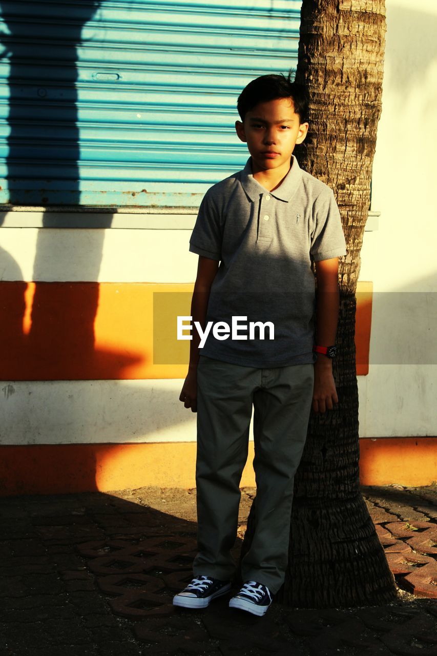 Full length portrait of boy standing by tree trunk on sidewalk